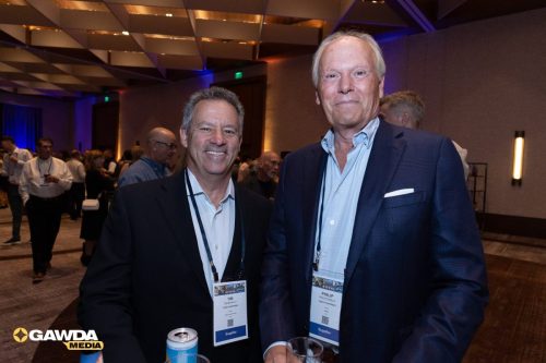 Two men in suits posing at a conference, wearing lanyards with badges and a crowded background.
