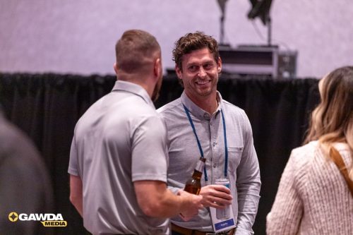 Two men converse at a conference, one holding a beer and the other a can, both wearing name badge lanyards, with GAWDA MEDIA logo in the corner.