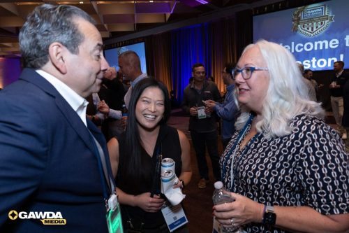 Networking reception at a conference with three people foreground: a man in a navy suit left, a smiling woman in black holding drinks center, and a woman with white hair and blue glasses on the right; badges and lanyards visible; GAWDA Media logo in the bottom-left corner.