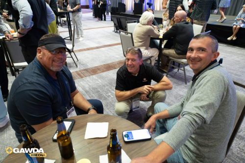Three men sit around a round table at a conference, smiling, with beer bottles and phones on the table and attendee badges visible on lanyards.