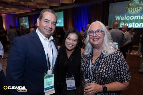 Three smiling conference attendees pose for a group photo, wearing badges, with a GAWDA Media logo and a 'Welcome to Boston' screen in the background.