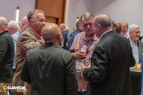 Group of men in business casual attire converse at a conference reception, holding drinks and wearing name badges.