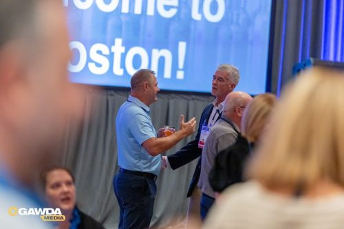 Two men in a conference setting converse, one holding a drink, with a large blue screen showing 'Boston' in the background.