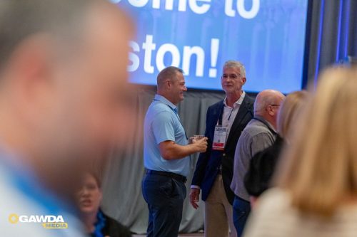 Two men in a conference setting chat, one in a light blue polo and the other in a dark blazer with a badge, with a large blue screen behind and blurred attendees in the foreground.