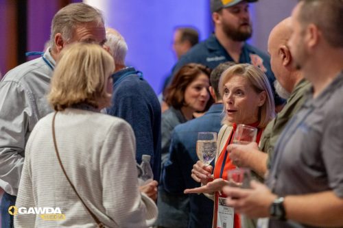 Woman in orange top speaks with a man, holding a wine glass at a crowded networking event; attendees mingle in the background.