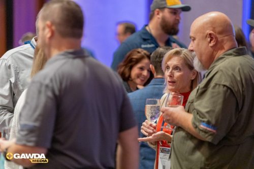 Two attendees converse at a social event, woman in red with a wine glass talking to a bald man in a green shirt; badge visible.