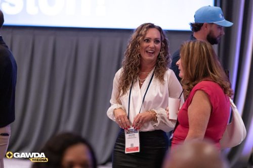 Two women in conversation at a conference: one with curly hair in a white blouse and name badge, the other in a pink top holding a cup of drink, on a stage backdrop with curtains behind them.