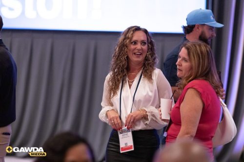 Two women chat at a conference: one in a white blouse with a lanyard, the other in pink holding a cup with a straw; a man in a blue cap stands behind them.