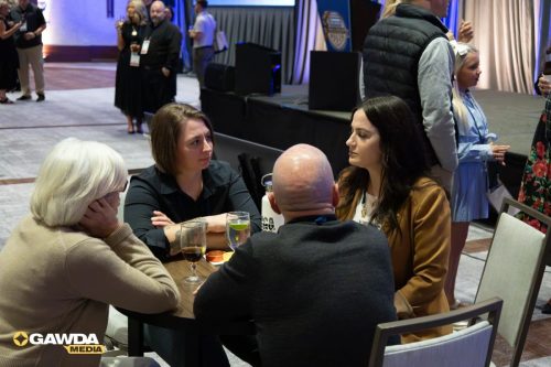 Four adults sit around a round table at an event, chatting with drinks on the table and a stage in the background.