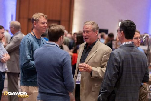 Group of men in business casual and suits chatting at a conference reception; one holds a drink.