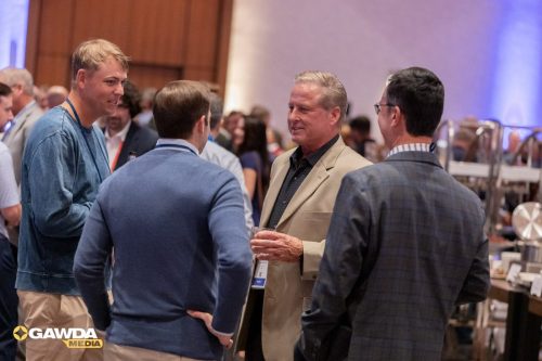 Four men in business casual attire chat at a crowded conference, one holding a glass.