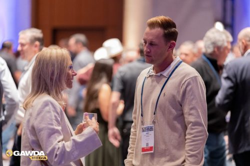 Two adults in a networking event chat: a blond woman with glasses holds a drink while speaking to a man with a name badge, with a crowded venue in the background (GAWDA Media logo bottom-left).