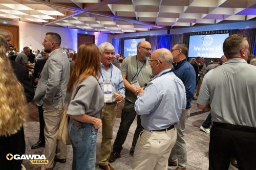 Group of professionals networking at a large conference hall, with badge lanyards and two screens reading 'Welcome to Boston!' in the background.
