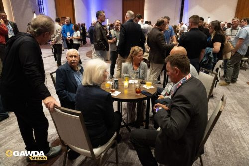 Networking event with professionals seated around a round table, drinks and a candle in the center, logo visible in corner, and people chatting in the background.