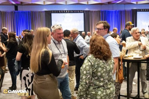 Crowd of professionals mingling at a conference reception, with high tables and blue/purple stage lighting in a large hall.
