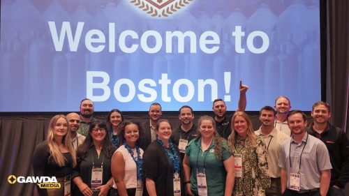 Group of attendees posing on stage in front of a 'Welcome to Boston!' screen at a conference or event badge visible on many people