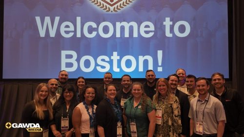 Group of professionals posing for a conference photo in front of a large screen that reads 'Welcome to Boston!', with a GAWDA Media logo in the bottom left.