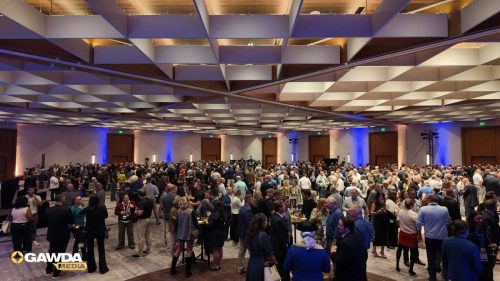 Large conference hall filled with people mingling at a networking event, geometric ceiling panels and blue-accented walls visible.