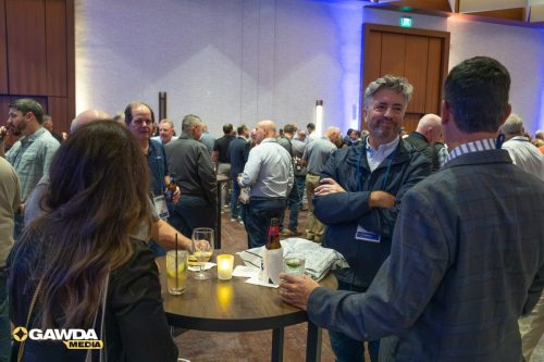 Networking event with people chatting around a high table, holding drinks in a conference hall; GAWDA MEDIA logo bottom left.