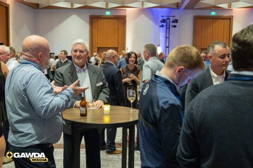 A group of professionals mingling around a high round table with drinks at a reception, candle lit, in a conference hall.