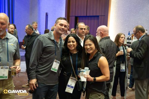 Three people in the foreground pose for a photo at a conference reception, wearing lanyards, while others mingle in the background.