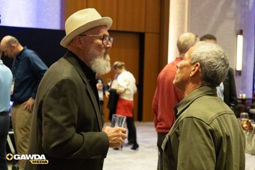 Two older men chat at a social event; one wears a light straw hat and glasses, holding a glass in his hand.