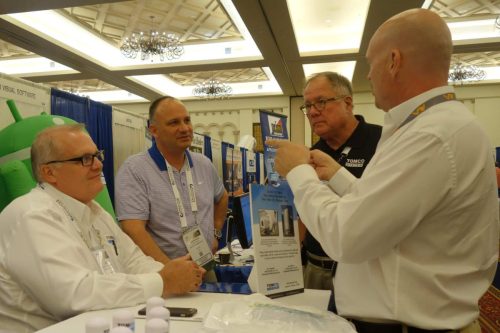 Three men at a trade show booth talking, with display boards and brochures in a ballroom setting.