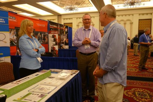 Woman in a light blue shirt stands behind a table of brochures at a trade show booth, talking to two men.