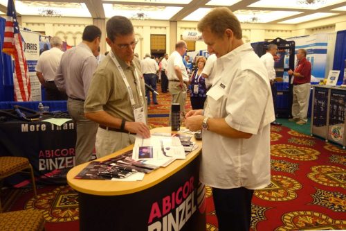 Two men at an ABICOR BINZEL booth examining brochures, with an American flag and other exhibition booths in the background.