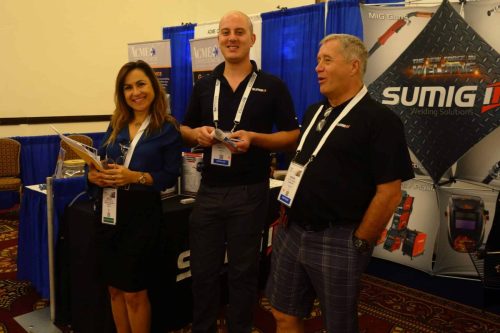 Three smiling attendees stand at a trade show booth with Sumig welding banners, each wearing event lanyards and holding brochures and samples.