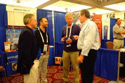 Group of men in a trade show booth, blue curtains, discussing welding products.