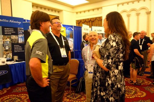 People converse at a trade show booth in a conference hall, with blue display banners and attendees wearing name badges.