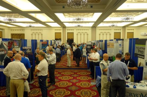 Busy convention hall with rows of blue exhibitor booths and attendees networking at a trade show event behind them, ornate ceiling visible above.