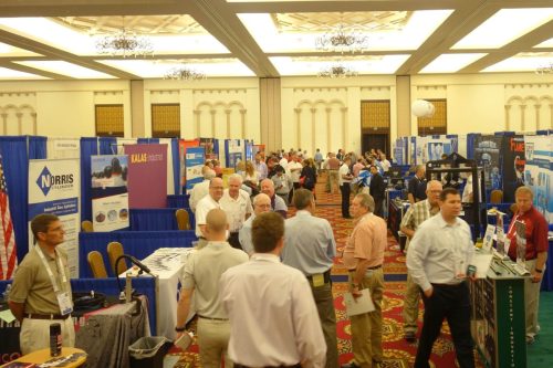 Busy trade show hall with numerous company booths and attendees browsing displays in a banquet-style venue.