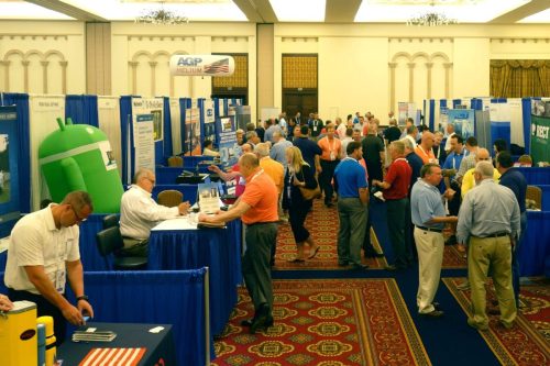 Busy trade show floor in a hotel ballroom with blue booth drapes and attendees talking at exhibitor tables.