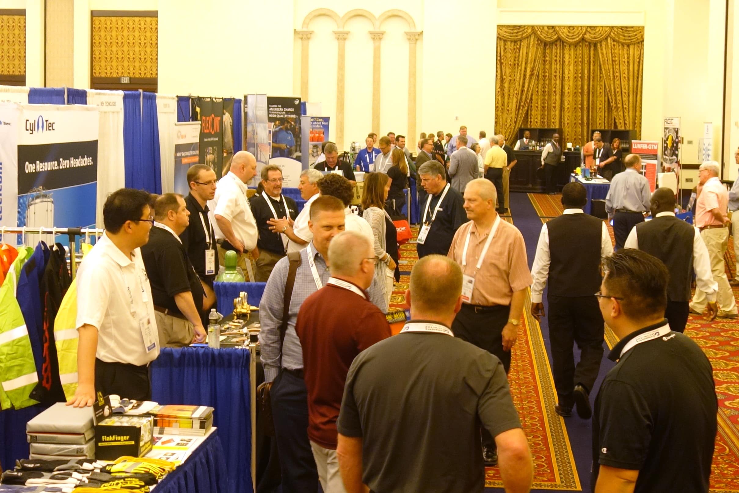 Busy trade show hall with attendees walking between vendor booths and blue-curtained displays, ornate carpet visible throughout.