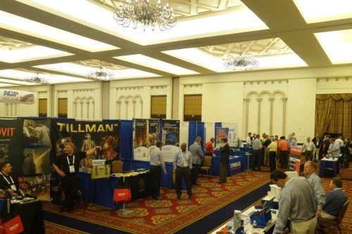 Trade show exhibition hall with multiple vendor booths, banners (Tillman, etc.), and attendees mingling under ornate chandeliers in a grand ballroom setting.