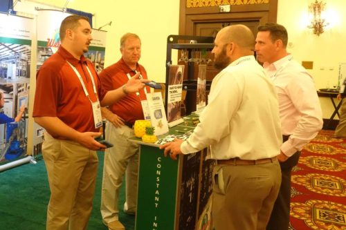 Four men in polo shirts talk at a trade show booth with brochures and product displays visible in the background.