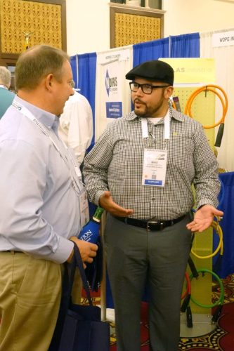 Two men at a trade show booth in conversation; left man in light blue shirt holding a navy bag, right man wearing a black cap, glasses, and a supplier badge with a checkered shirt.