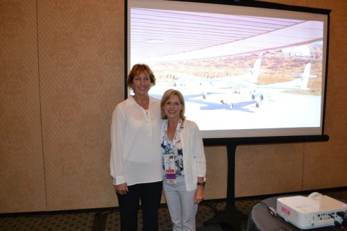 Two women pose for a photo in a conference room with a projector screen displaying a desert airstrip and small airplanes in the background, both smiling.