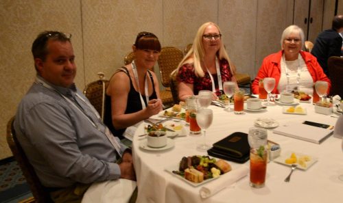 Four people seated at a banquet table during a meal, with plates of food and tall drinks; a man on the left and three women on the right.