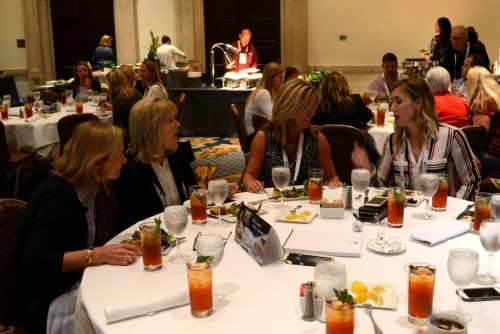 People seated at round tables in a banquet hall during a dinner event, with drinks and plates of food in front of them.