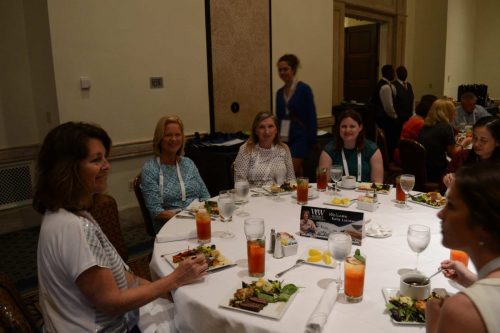 Group of people seated around a round banquet table with plates of salad, drinks, and cutlery, chatting as they eat.