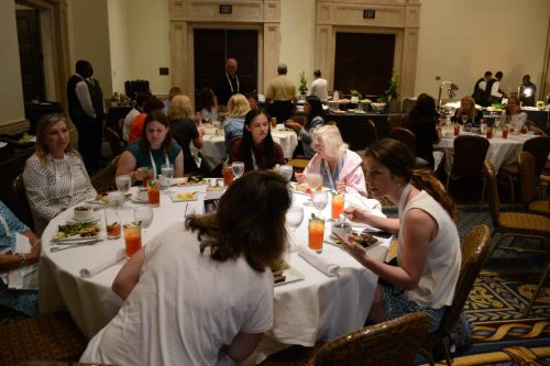 People seated at round banquet tables eating and chatting in a formal dining room with a buffet in the background.