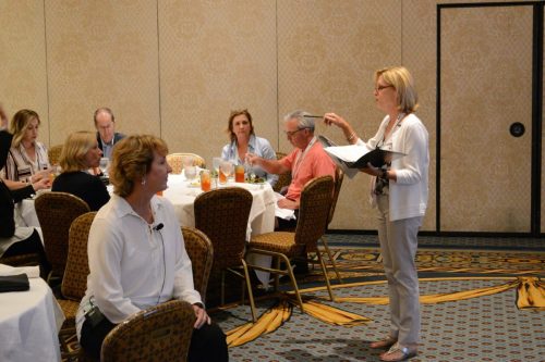 Female presenter in a white blazer speaks to a seated audience at a banquet-style meeting room.
