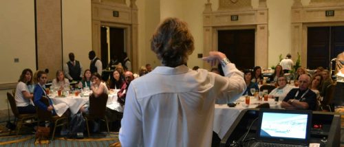 Speaker with short hair in a white shirt addresses a seated banquet audience in a large ornate room.