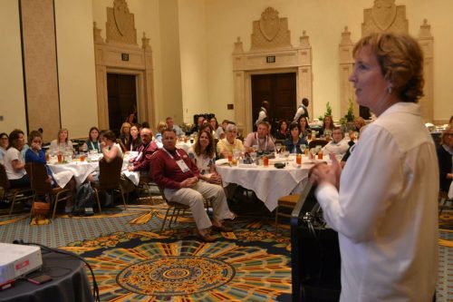 Speaker at a podium addressing a banquet hall full of attendees seated at round tables with drinks and plates in front of them.