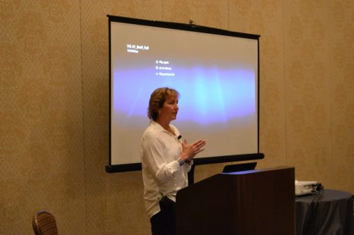 Female presenter in a white blouse at a lectern, speaking in front of a projection screen in a beige-patterned room.