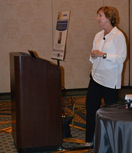 Side view of a woman in a white blouse speaking at a wooden podium with a microphone, a poster board in the background.