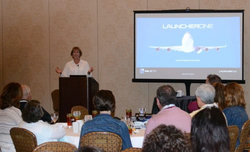 Female presenter at a lectern giving a talk, with a large projector slide showing an airplane and the word 'LAUNCHERONE' in a conference room.
